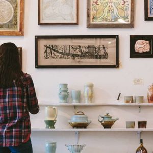 A woman in a plaid shirt admiring framed artwork on a wall, with shelves below displaying pottery and ceramic pieces.