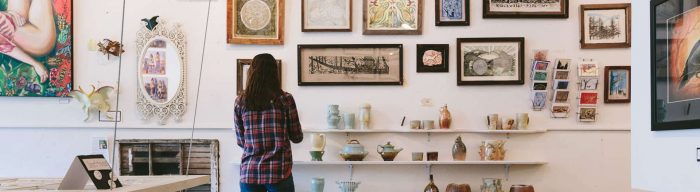 A woman in a plaid shirt admiring framed artwork on a wall, with shelves below displaying pottery and ceramic pieces.
