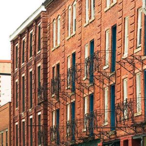 A row of historic red brick buildings with black fire escapes, alongside colorful storefronts and balconies on a tree-lined street.