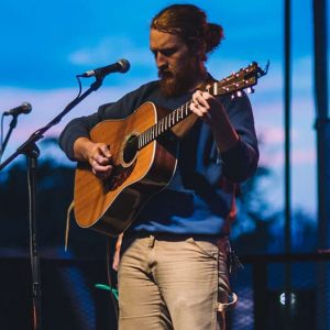 A band performing on an outdoor stage at dusk, featuring a guitarist, a vocalist with an acoustic guitar, and a musician playing a pedal steel guitar.