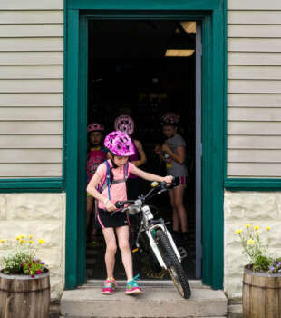 A child wearing a pink helmet walking a bicycle out of a doorway, with other children and bikes visible inside.