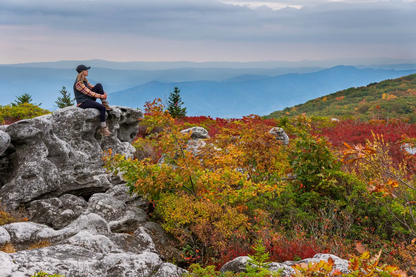 A woman in hiking gear sits atop a rugged rock formation, surrounded by vibrant fall foliage with layered mountain ridges fading into the distance under a cloudy sky.