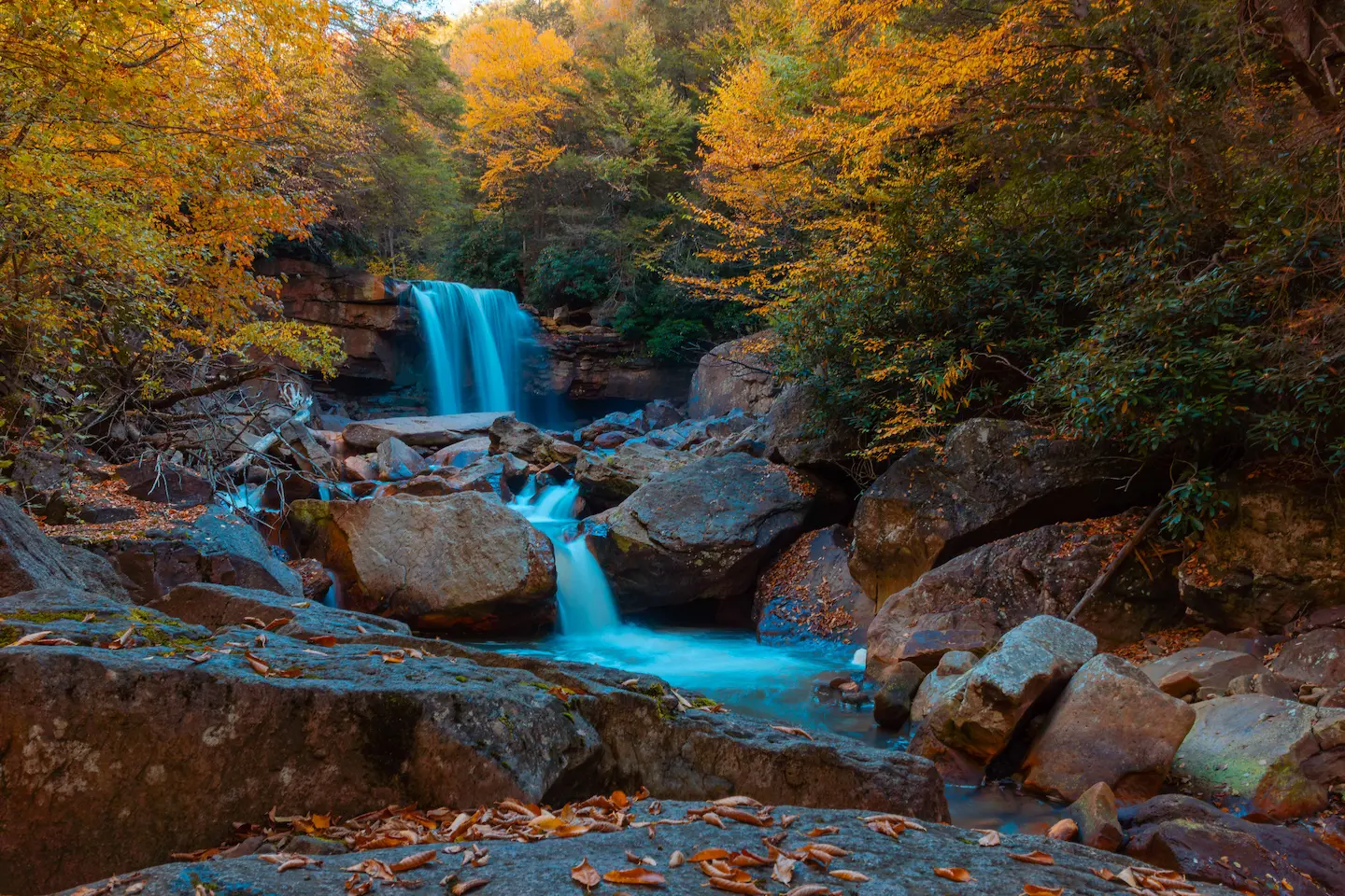 A scenic waterfall cascades over rocks into a clear blue pool, framed by autumn trees with golden and orange leaves in a dense forest.