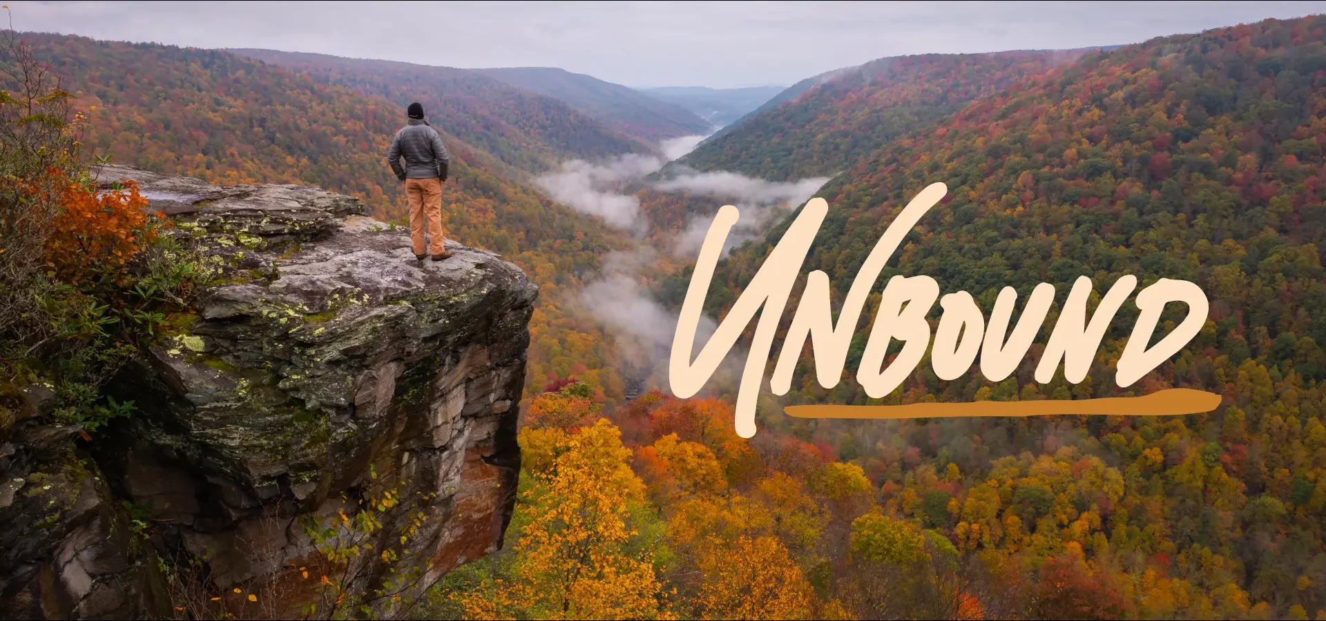 A person stands on the edge of a rocky cliff overlooking a vast, fog-filled valley surrounded by colorful autumn foliage, with the word “Unbound” written in bold script across the scene.